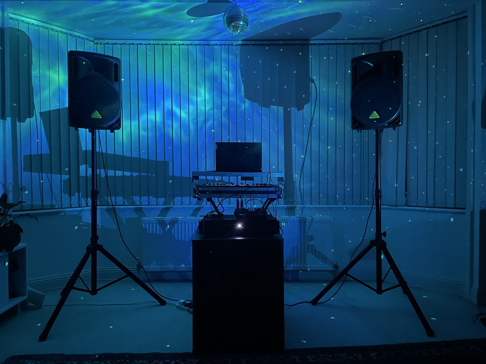 The PA system set up in a room: two speakers on tripod stands either side of a DJ controller and laptop, with a subwoofer beneath and a mirror ball overhead.